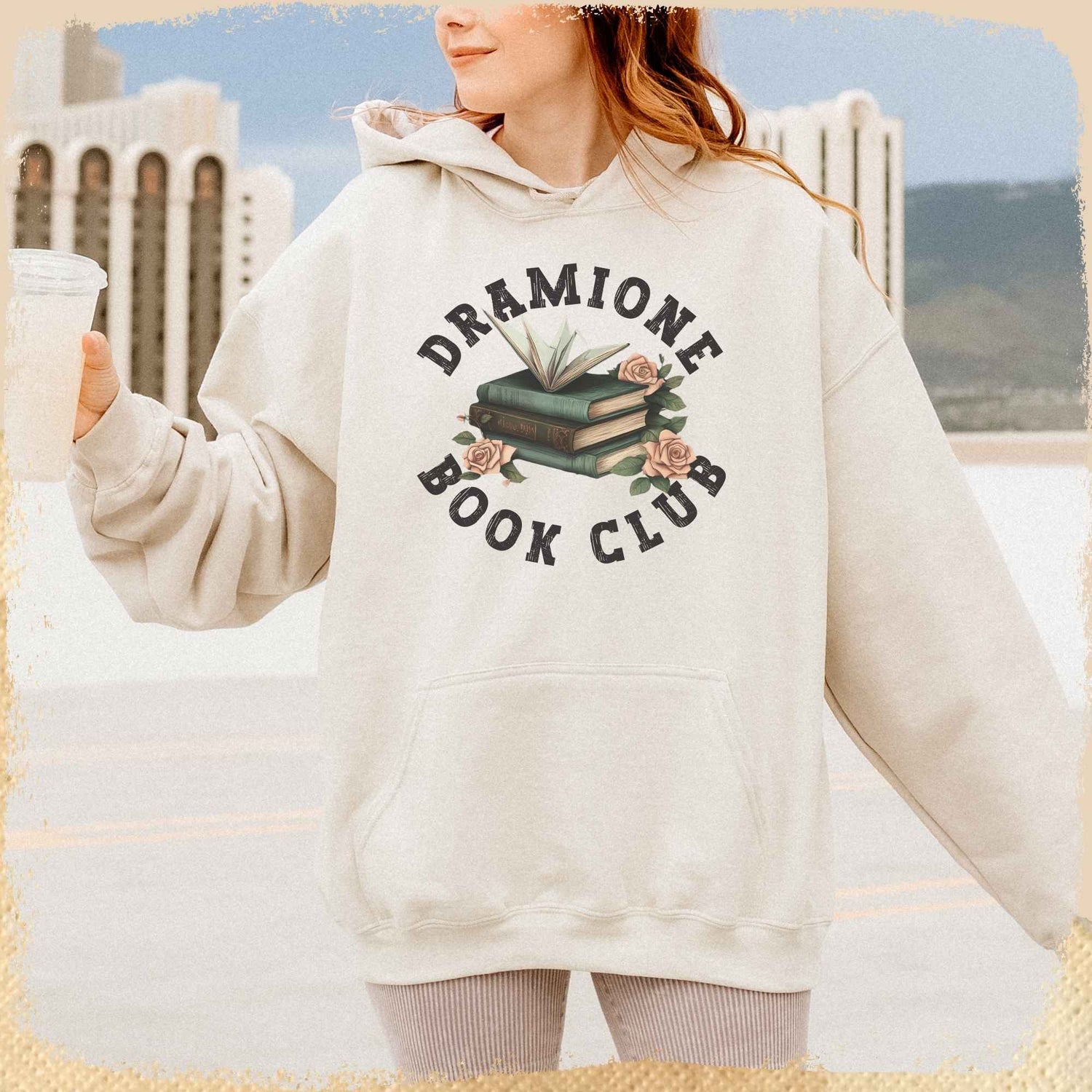 Dramione Hoodie getragen von einer jungen Frau. Der sandfarbene Oversizehoodie trägt in der Mitte ein Motiv von einem Bücherstapel auf dem ein Papierkranich sitzt. Rund angeordnet um das Motiv, ist in Collegeshrift der Schriftzug Dramione Book Club zu lesen.