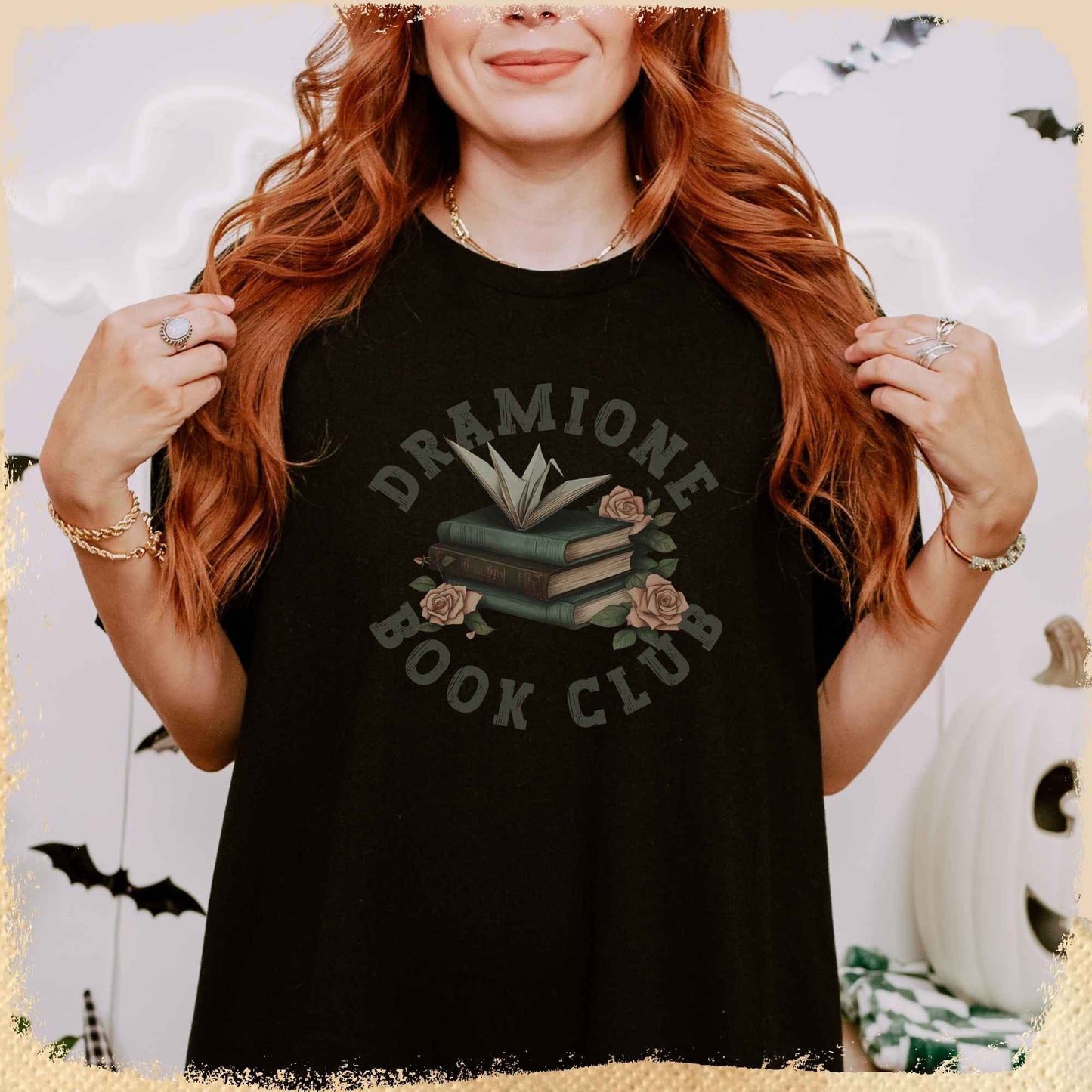 Eine Frau mit langen, roten Haaren trägt ein schwarzes T-Shirt mit der Aufschrift „Dramione Book Club“. In der Mitte des Shirts ist ein Stapel Bücher mit Rosen und einem aufgeklappten Buch als Dekor abgebildet. Im Hintergrund sind herbstliche und Halloween-Dekorationen wie Fledermäuse und ein weißer Kürbis zu sehen.