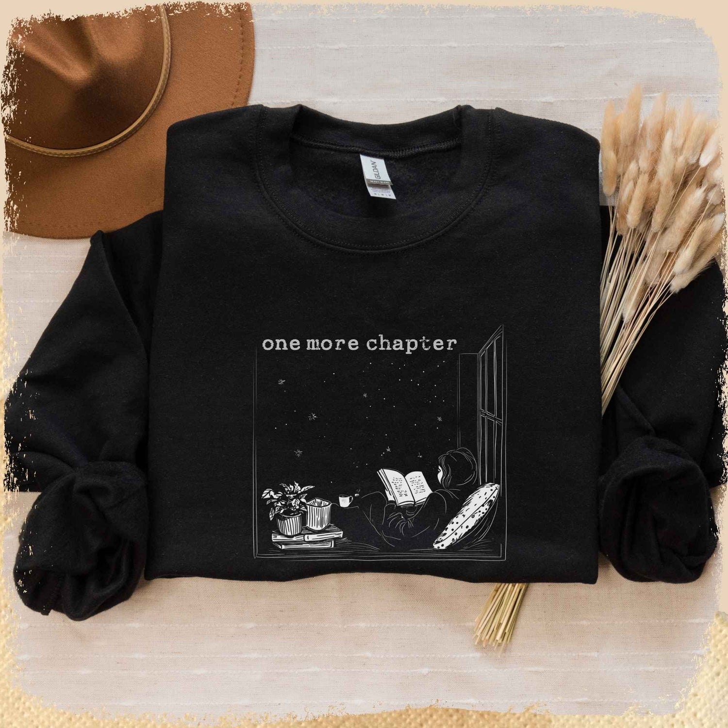 Man sieht ein schwarzes Book Sweatshirt mit einer weißen Skizze von einer Leserin, die auf einem Fensterbrett sitzt und in ein Buch vertieft ist. Draußen funkeln die Sterne. Über der Skizze steht "one more chapter".