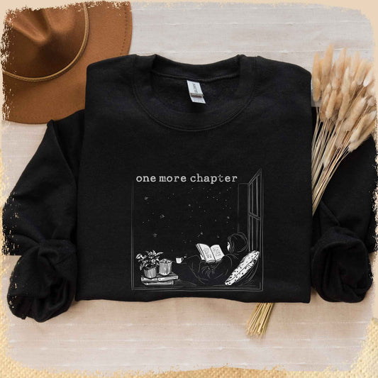 Man sieht ein schwarzes Book Sweatshirt mit einer weißen Skizze von einer Leserin, die auf einem Fensterbrett sitzt und in ein Buch vertieft ist. Draußen funkeln die Sterne. Über der Skizze steht "one more chapter".
