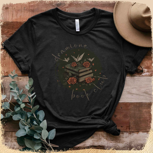 Ein schwarzes T-Shirt mit Dramione-Book-Club-Motiv in Flatlay-Perspektive. Papierkraniche fliegen über einen Bücherstapel, der von Rosen umgeben ist.