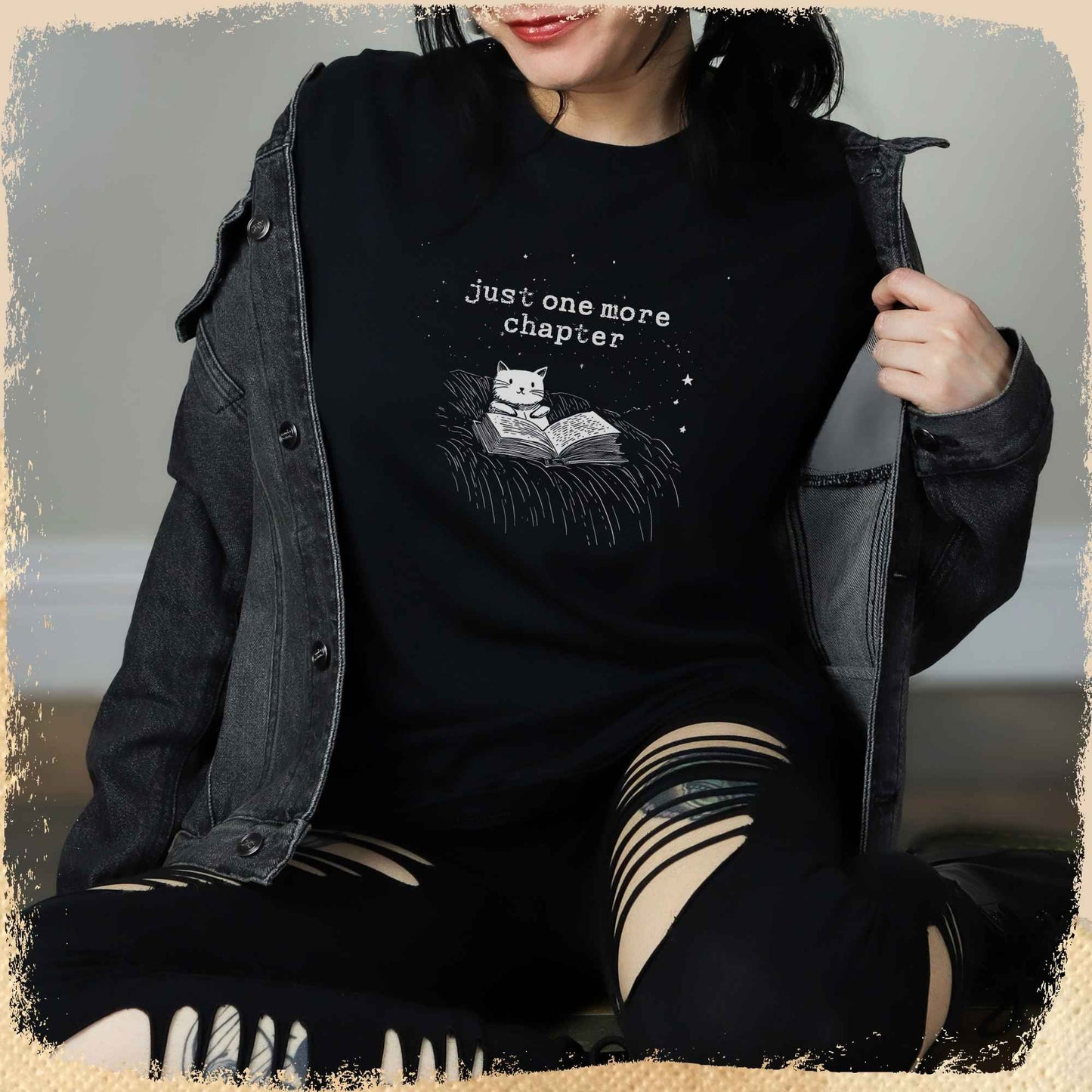Eine junge Frau trägt ein schwarzes T-Shirt mit einem minimalistischen Design: Eine gezeichnete Katze liest ein Buch unter einem Sternenhimmel, darüber steht der Schriftzug „just one more chapter“. Das Outfit wird mit einer dunklen Jacke und zerrissenen Jeans kombiniert.