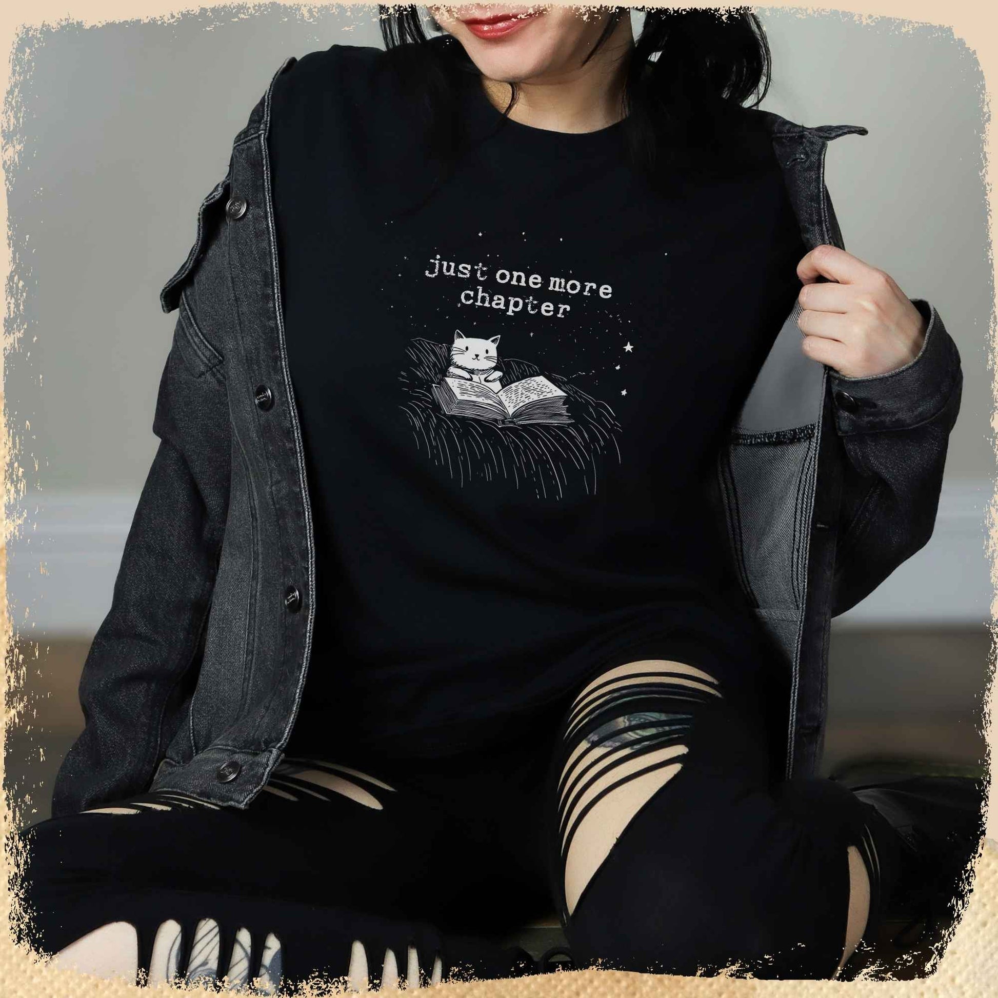 Eine junge Frau trägt ein schwarzes T-Shirt mit einem minimalistischen Design: Eine gezeichnete Katze liest ein Buch unter einem Sternenhimmel, darüber steht der Schriftzug „just one more chapter“. Das Outfit wird mit einer dunklen Jacke und zerrissenen Jeans kombiniert.