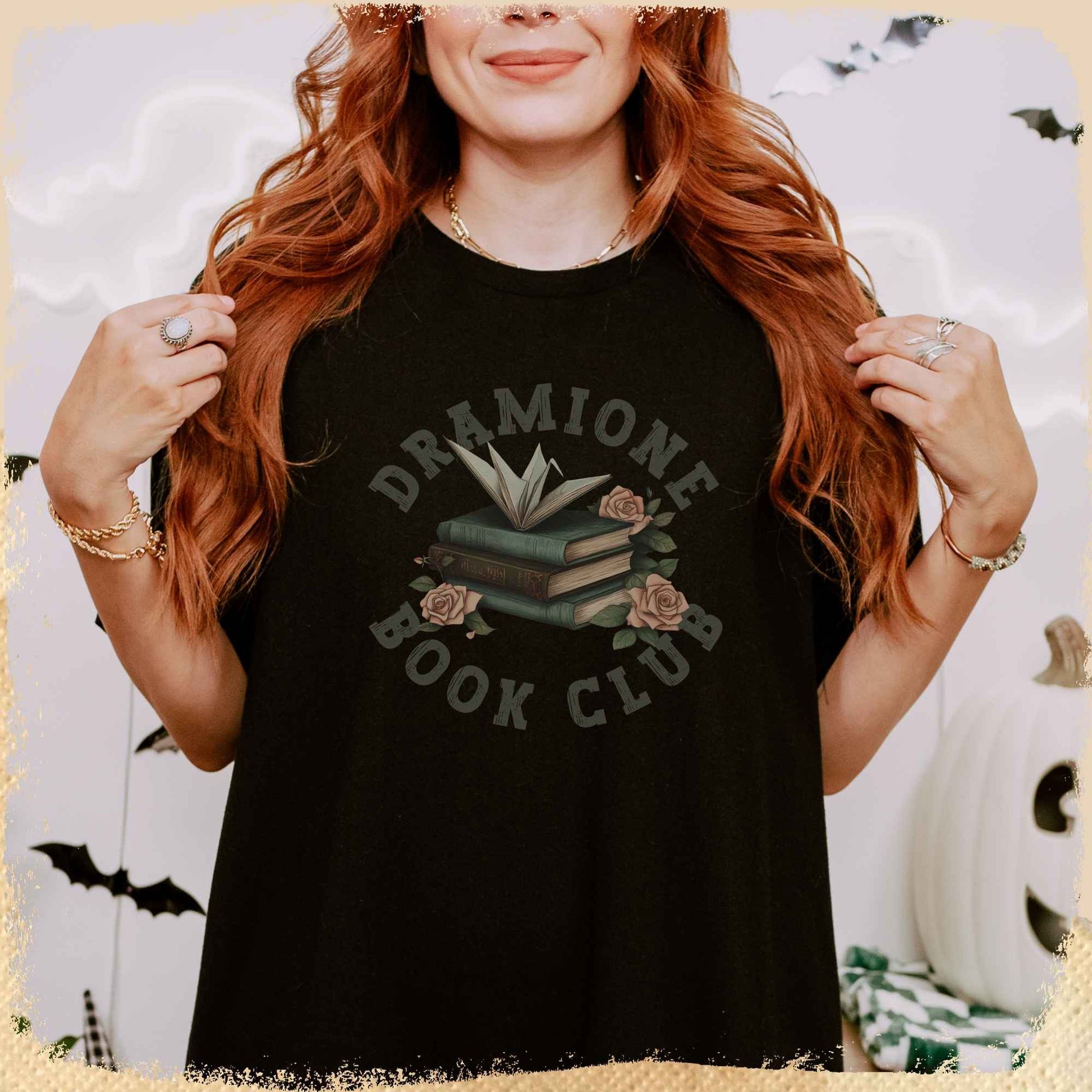 Eine Frau mit langen, roten Haaren trägt ein schwarzes T-Shirt mit der Aufschrift „Dramione Book Club“. In der Mitte des Shirts ist ein Stapel Bücher mit Rosen und einem aufgeklappten Buch als Dekor abgebildet. Im Hintergrund sind herbstliche und Halloween-Dekorationen wie Fledermäuse und ein weißer Kürbis zu sehen.