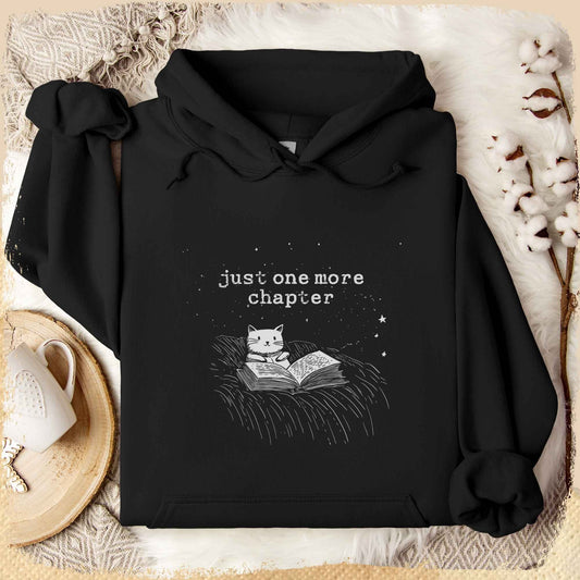Schwarzer Hoodie mit weißem Print: Eine gezeichnete Katze liegt gemütlich auf einer Decke und liest ein Buch unter einem sternenklaren Himmel. Darüber steht in verspielter Schrift „Just one more chapter“. Der Hoodie liegt dekorativ arrangiert auf einem weichen Untergrund, umgeben von Baumwollzweigen und einer Tasse Tee.